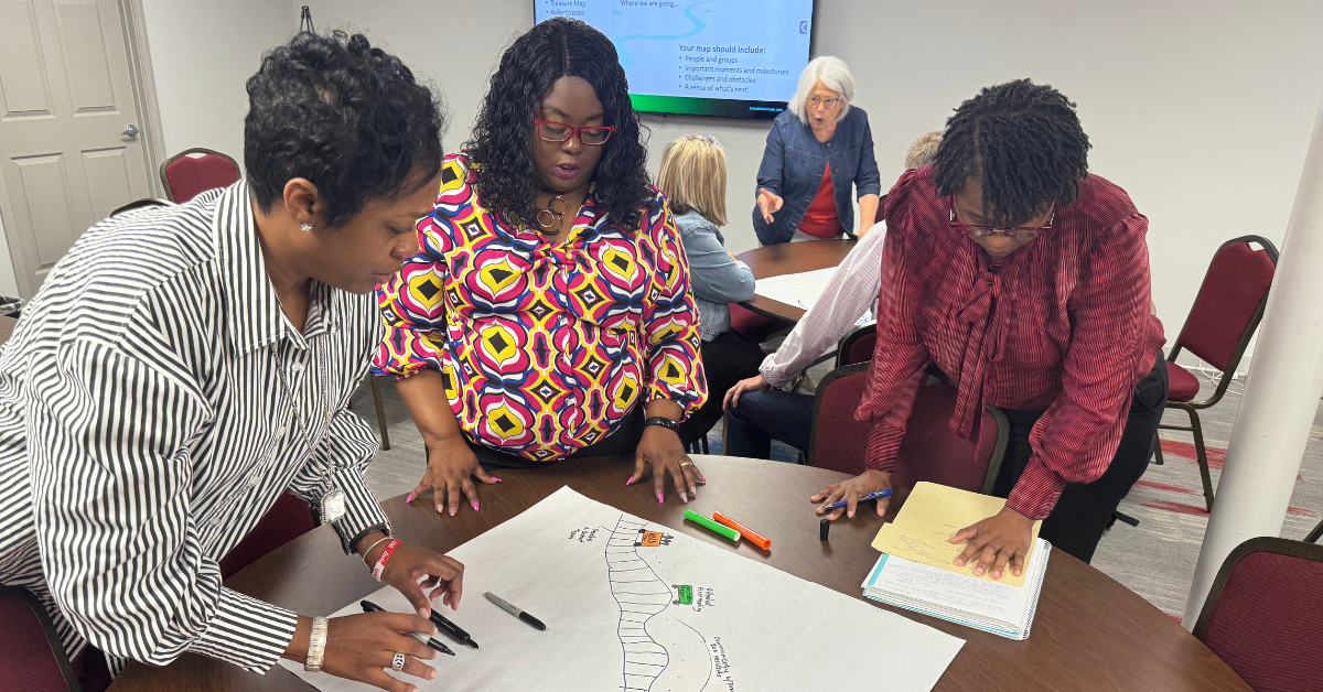 Three women are huddled around a table outlining their their school improvement journey using markers and anchor chart paper.